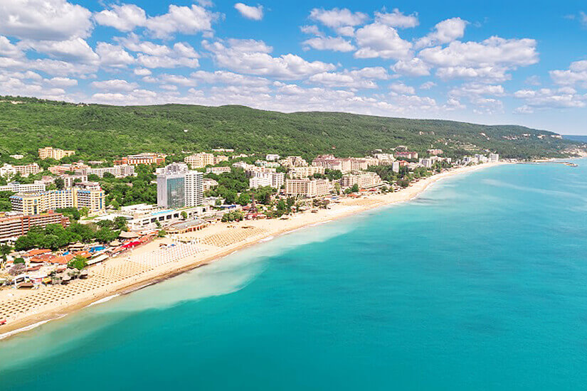 Aus der Luft betrachtet erstreckt sich ein breiter goldgelber Sandstrand entlang der türkisblauen Küste, gesäumt von zahlreichen Hotelanlagen und dichten, bewaldeten Hügeln unter einem Himmel mit Schäfchenwolken. Reiseziele August Goldstrand