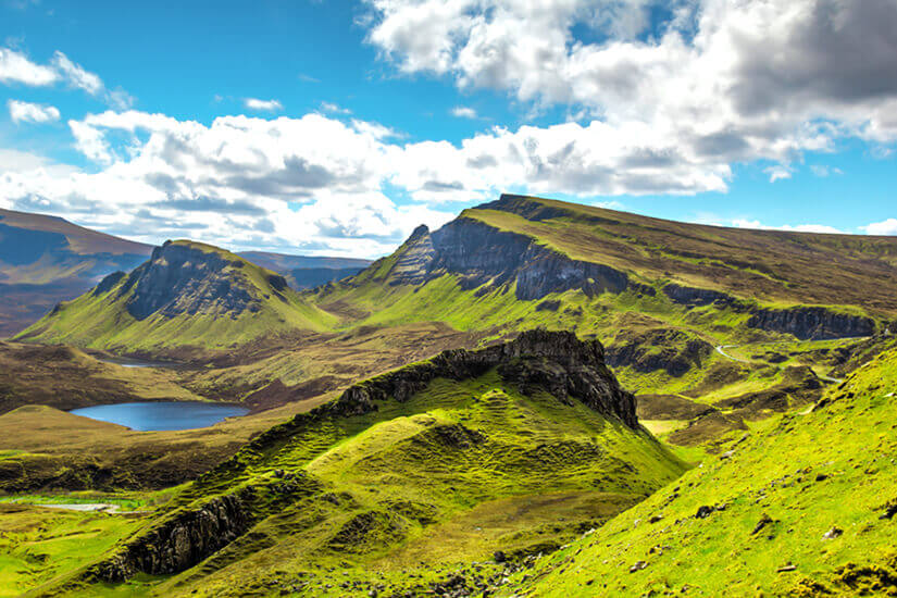 Weitläufig erstreckt sich eine dramatische Landschaft aus saftig grünen Hügeln, schroffen Felsformationen und einem kleinen Bergsee unter einem lebhaften Wolkenhimmel. Reiseziele August Schottland