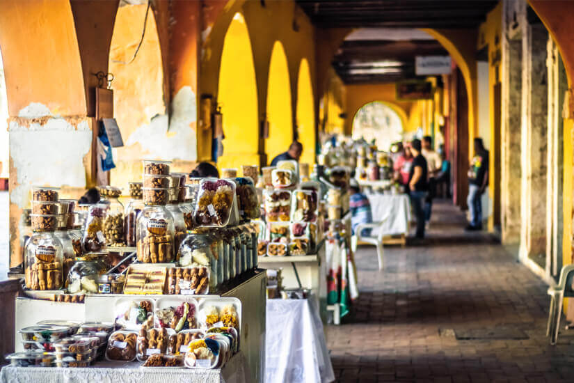 Entlang eines gelben Arkadenganges reihen sich Marktstände mit Glasbehältern voller traditioneller Süßigkeiten und Snacks, während im Hintergrund Passanten durch die kolonialen Gewölbe schlendern. Reiseziele August Kolumbien