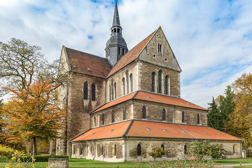 Zu erkennen ist eine große Klosterkirche aus Naturstein mit einem hohen Turm und roten Ziegeldächern. Das Bauwerk ist von Rasenflächen und Bäumen umgeben, während sich darüber ein teilweise bewölkter Himmel erstreckt. Niedersachsen Riddagshausen
