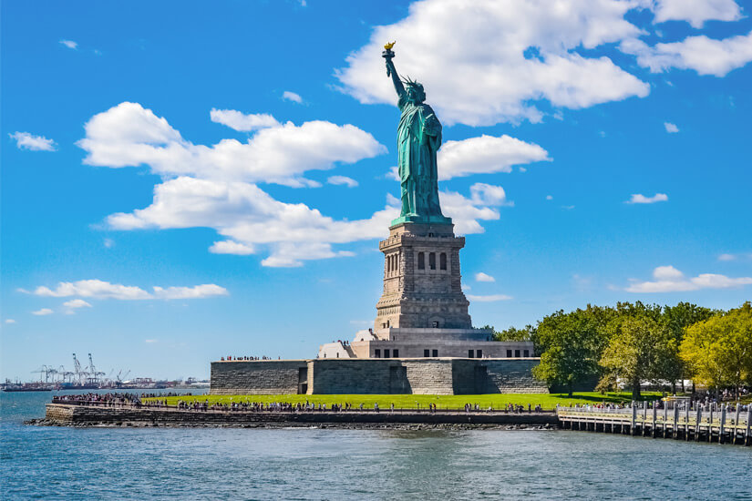 Majestätisch erhebt sich die weltberühmte Freiheitsstatue mit ihrer erhobenen Fackel auf einem steinernen Sockel über Liberty Island, umrahmt von einem lebhaften Wolkenhimmel und dem glitzernden Wasser des Hafens von New York. Reiseziele August USA