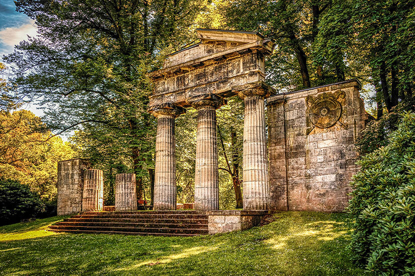 Zu erkennen ist ein monumentales Bauwerk im Stil eines antiken Tempels mit hohen Säulen und einem Giebeldach, eingebettet in eine weitläufige Parkanlage. Umgeben wird die Ruine von alten Bäumen, gepflegten Rasenflächen und dichtem Grün. Niedersachsen Buergerpark