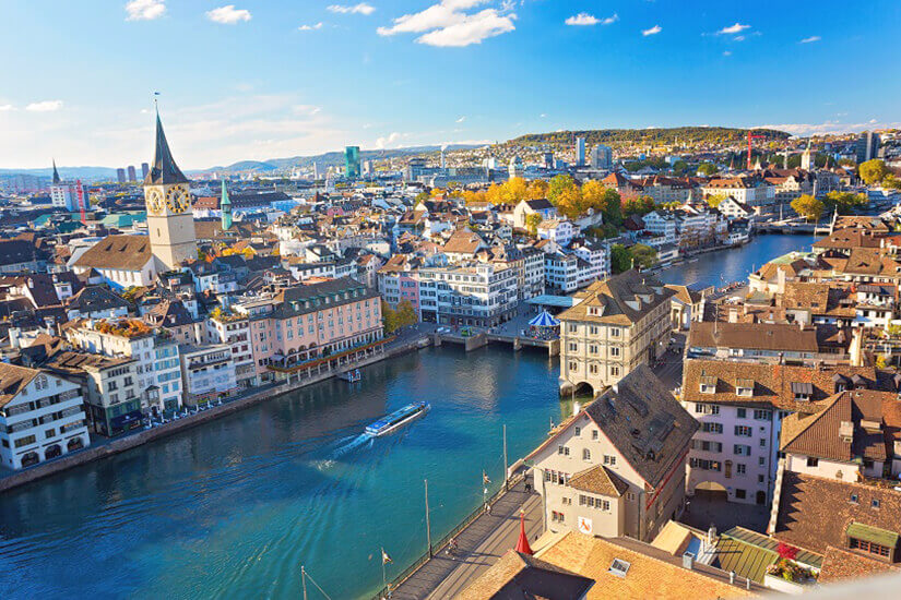 Vom erhöhten Standpunkt aus erstreckt sich ein malerischer Blick über die Altstadt mit ihren historischen Häuserdächern, den markanten Kirchtürmen und einem Boot auf dem türkisblauen Fluss unter einem sonnigen Himmel mit Wolken. Reiseziele August Schweiz