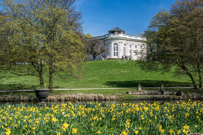 Zu erkennen ist ein elegantes Schloss auf einer Anhöhe inmitten einer weitläufigen Parkanlage. Im Vordergrund blühen zahlreiche gelbe Blumen an einem Teich, während hohe Bäume das Gebäude auf beiden Seiten einrahmen. Niedersachsen Schloss Richmond