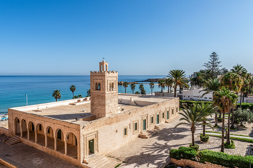 Eine Weitwinkelaufnahme der historischen Festung Ribat von Monastir in Tunesien aus der Vogelperspektive. Das sandfarbene Steinbauwerk verfügt über einen quadratischen Minarett-Turm und eine überdachte Säulengalerie im Erdgeschoss. Das Gebäude liegt direkt an der Küste, wo das tiefblaue Mittelmeer unter einem wolkenlosen, hellblauen Himmel an den Strand trifft. Rechts von der Festung erstreckt sich eine Parkanlage mit hohen Dattelpalmen und gepflegten Grünflächen. All Inclusive Urlaubsziele Monastir