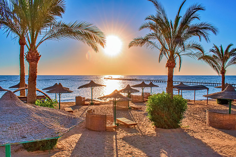Ein sonnenverwöhnter Sandstrand auf der Sinai-Halbinsel im warmen Gegenlicht der tiefstehenden Sonne. Auf dem feinen Sand stehen mehrere strohgedeckte Sonnenschirme, hölzerne Sonnenliegen und geflochtene Windschutze. Schlanke Palmen werfen lange Schatten über den Strand. Im Hintergrund glitzert das ruhige Meer, in das ein langer Steg hineinragt, unter einem wolkenlosen, leuchtend blauen Himmel. All Inclusive Urlaubsziele Sinai