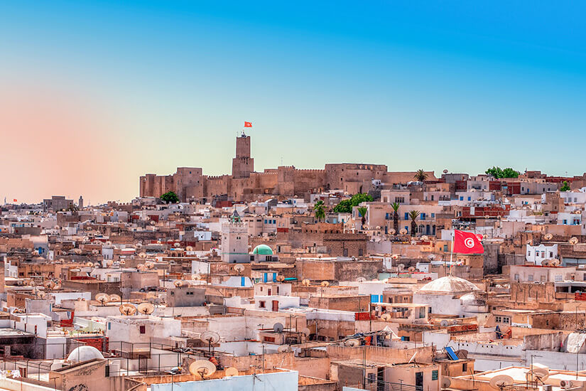 Ein Panoramablick über die Dächer der Medina von Sousse in Tunesien. Dicht aneinandergebaut prägen helle, flache Gebäude mit zahlreichen Satellitenschüsseln das Stadtbild. Im Hintergrund erhebt sich auf einer Anhöhe die beeindruckende, mauerumgebene Festungsanlage des Ribat mit ihrem hohen Wachturm, auf dem die tunesische Flagge weht. Der Himmel verläuft von einem sanften Abendrot am Horizont zu einem klaren Blau oben. All Inclusive Urlaubsziele Sousse