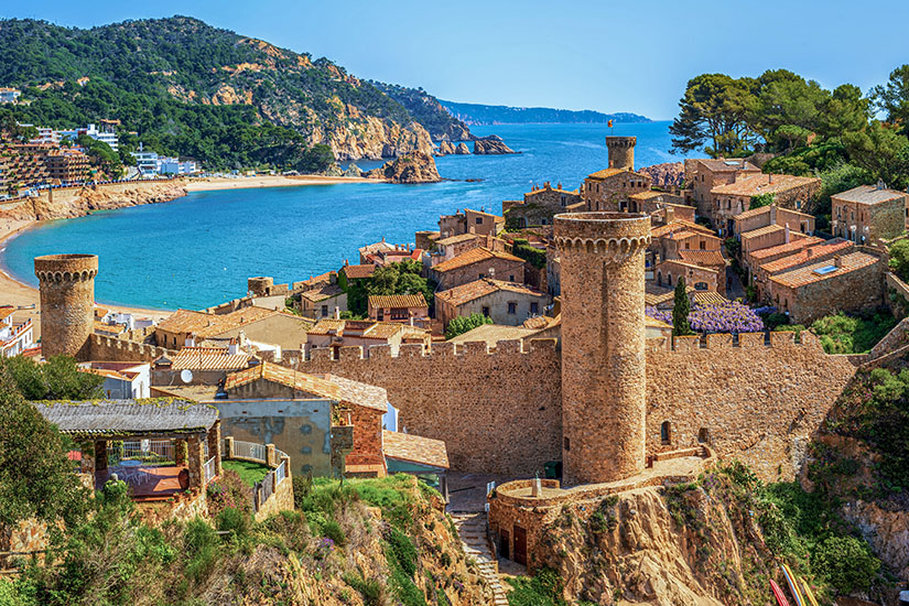 Eine erhöhte Ansicht der mittelalterlichen Festung und Altstadt von Tossa de Mar in Katalonien. Im Vordergrund erstrecken sich massive, braune Steinmauern mit rund ausgeprägten Wehrtürmen. Dahinter schmiegen sich Häuser mit roten Ziegeldächern an den Hang. Die Festung liegt unmittelbar an einer geschwungenen Meeresbucht mit türkisblauem Wasser, goldenem Sandstrand und dicht bewachsenen, grünen Hügeln im Hintergrund unter strahlend blauem Himmel. All Inclusive Urlaubsziele Katalonien