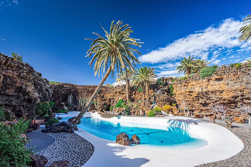 Ein geschwungener, leuchtend türkisblauer Pool mit strahlend weißer Umrandung im Jameos del Agua auf Lanzarote. Das Baddomizil liegt eingebettet in eine tief gelegene Senke aus dunklem, felsigem Vulkangestein. Große, grüne Palmen wachsen direkt am Beckenrand und an den felsigen Terassen empor, während darüber ein intensiver, blauer Himmel mit leichten, weißen Schleierwolken erstrahlt. All Inclusive Urlaubsziele Lanzarote