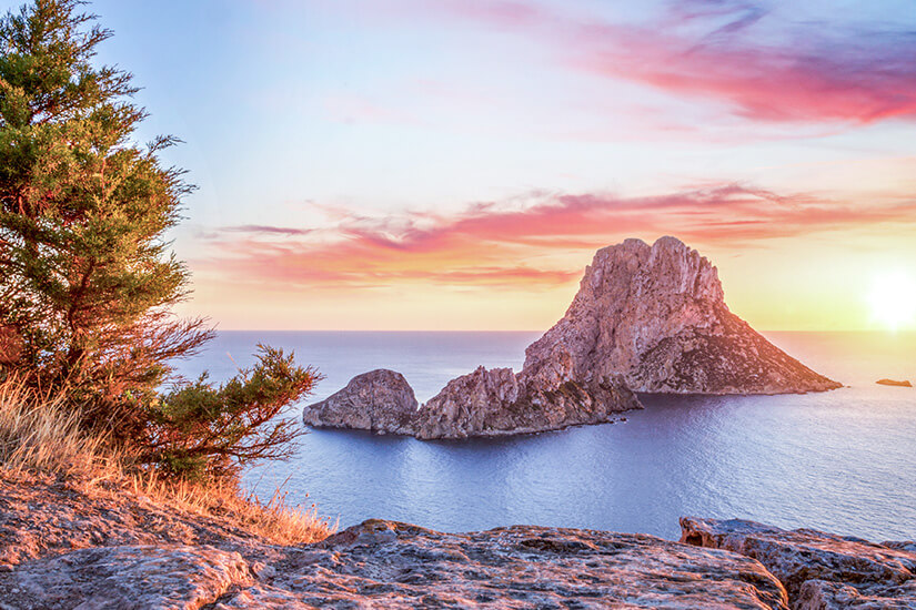 Ein spektakulärer Sonnenuntergang an der Küste von Ibiza mit Blick auf die magische Felseninsel Es Vedrà. Im Vordergrund liegt ein steiniges Plateau mit einem grünen Wacholderstrauch auf der linken Seite. Die Felseninsel ragt erhaben aus dem tiefblauen Meer hervor, während der Himmel in leuchtenden Rosa-, Orange- und Violetttönen erstrahlt und die tiefstehende Sonne ein warmes Licht über die Szenerie wirft. All Inclusive Urlaubsziele Ibiza