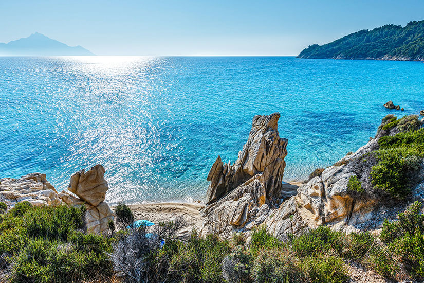 Ein Blick von oben auf eine idyllische Bucht auf Chalkidiki mit kristallklarem, türkisblauem Meer, das im Sonnenlicht glitzert. Im Vordergrund prägen helle, markant geformte Felsformationen und grüne Büsche das Ufer. Ein kleiner, versteckter Sandstrand liegt geschützt zwischen den Felsen. Im Hintergrund hebt sich im dunstigen Sonnenlicht die Silhouette eines Berges am Horizont ab. All Inclusive Urlaubsziele Chalkidiki
