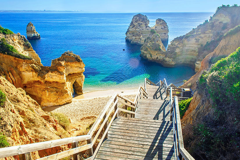 Eine Holztreppe führt steil hinab zu einer versteckten Bucht an der Ponta da Piedade an der Algarve. Der feine Sandstrand wird von spektakulären, golden leuchtenden Sandsteinfelsen und Felsformationen im tiefblauen Atlantik eingerahmt. Das seichte Wasser in Strandnähe schimmert in klarem Türkis unter einem sonnigen, strahlend blauen Himmel. All Inclusive Urlaubsziele Algarve