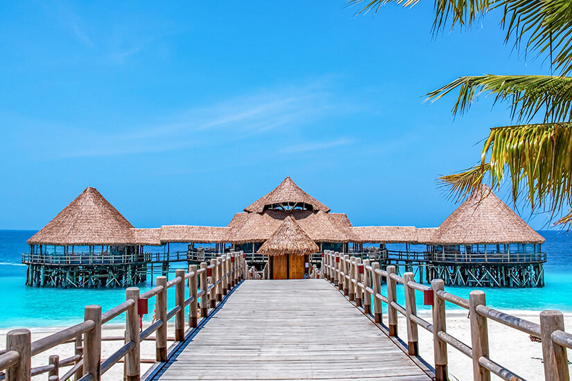 Ein langer Holztsteg mit Holzgeländer führt direkt vom weißen Sandstrand auf das leuchtend türkisblaue Wasser des Indischen Ozeans auf Sansibar zu. Am Ende des Stegs steht eine auf Pfählen erbaute Resort-Anlage mit reetgedeckten Pavillons und Spitzdächern. Rechts oben ragen grüne Palmenwedel ins Bild, während im Hintergrund ein strahlend blauer Himmel zu sehen ist. All Inclusive Urlaubsziele Sansibar