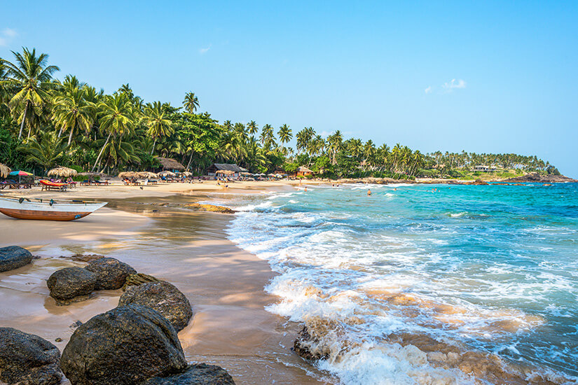 Ein traumhafter Tropenstrand auf Sri Lanka im hellen Tageslicht. Im Vordergrund spülen sanfte, weiße Schaumkronen des türkisblauen Indischen Ozeans über den goldenen Sand und dunkle Felssteine am Ufer. Links liegt ein schmales Holzboot im Sand. Entlang des geschwungenen Küstenstreifens erstreckt sich eine dichte Kulisse aus dichten, hohen Kokospalmen und kleinen Holzhütten unter einem klaren, blauen Himmel. All Inclusive Urlaubsziele Sri Lanka