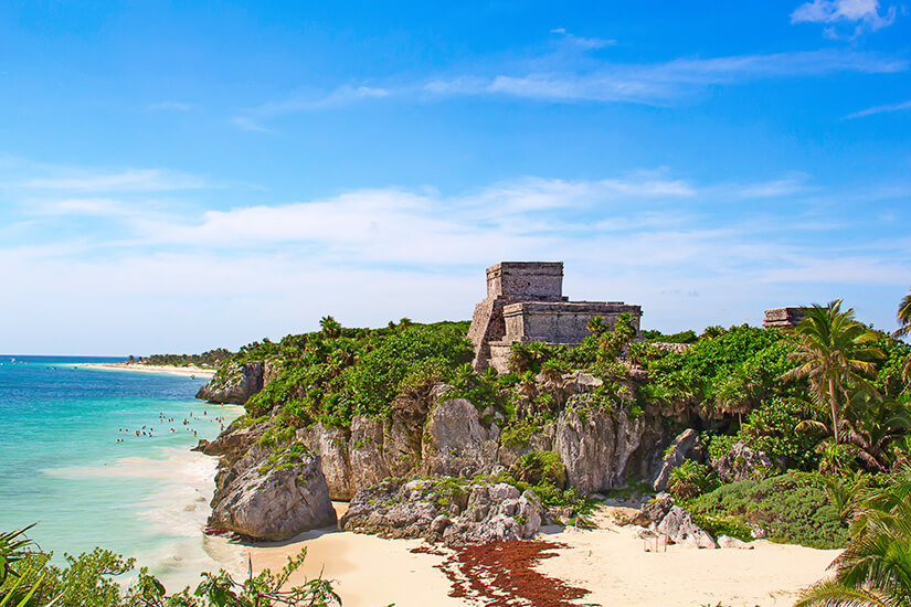 Die historischen Maya-Ruinen von Tulum thronen auf einer steil abfallenden, felsigen Klippe an der Riviera Maya in Mexiko. Das antike Steinbauwerk ist von üppig grüner, tropischer Vegetation und Palmen umgeben. Direkt unterhalb der Klippe erstreckt sich ein weißer Sandstrand, an den das schimmernde, türkisblaue Karibische Meer unter einem strahlend blauen Himmel mit feinen Schleierwolken grenzt. All Inclusive Urlaubsziele Riviera Maya