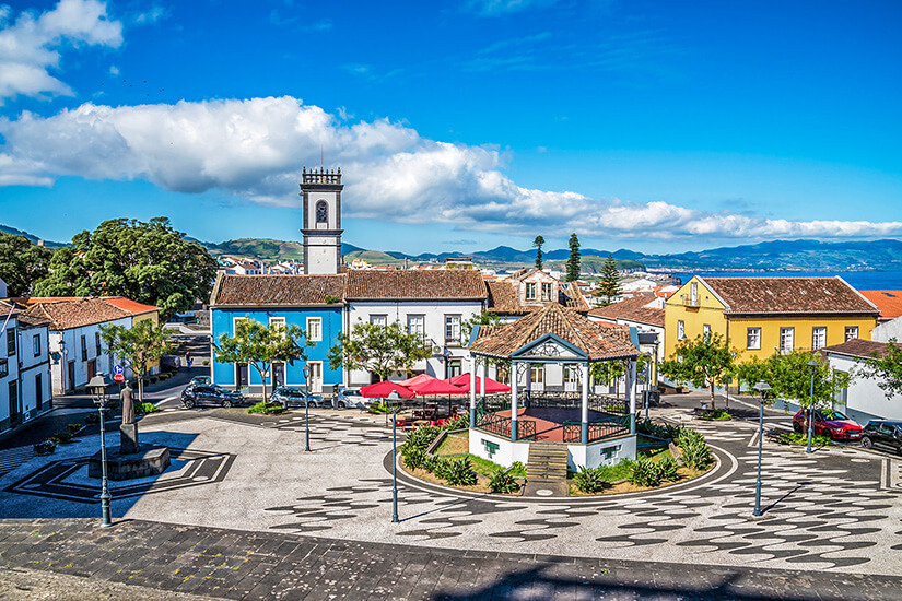 Ein farbenfroher Platz mit Kirchturm und traditionellem Pavillon. Blaue, gelbe und weiße Gebäude umrahmen das gepflasterte Zentrum. Azoreanisches Dorfleben in seiner schönsten Form. Azoren Portugal Kirche