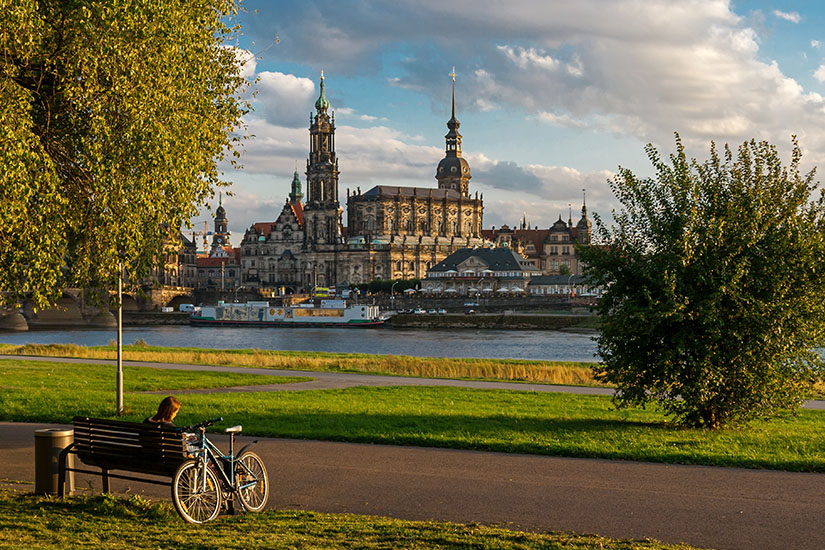 Zu erkennen ist ein Flussufer mit einem Radweg und einer Bank, neben der ein Fahrrad abgestellt ist. Auf der gegenüberliegenden Seite des Flusses erhebt sich eine historische Stadtsilhouette mit mehreren markanten Kirchen und Türmen unter einem teils bewölkten Himmel. Radwege Deutschland Elberadweg
