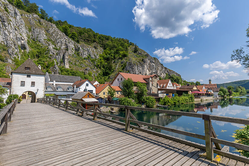 Zu erkennen ist eine historische Ortschaft an einem ruhigen Fluss, die von steilen Felswänden und bewaldeten Hängen umgeben ist. Im Vordergrund führt eine hölzerne Brücke auf ein Torhaus zu, während sich entlang des Ufers Häuser mit roten Ziegeldächern aneinanderreihen. Radwege Deutschland Altmuehltal