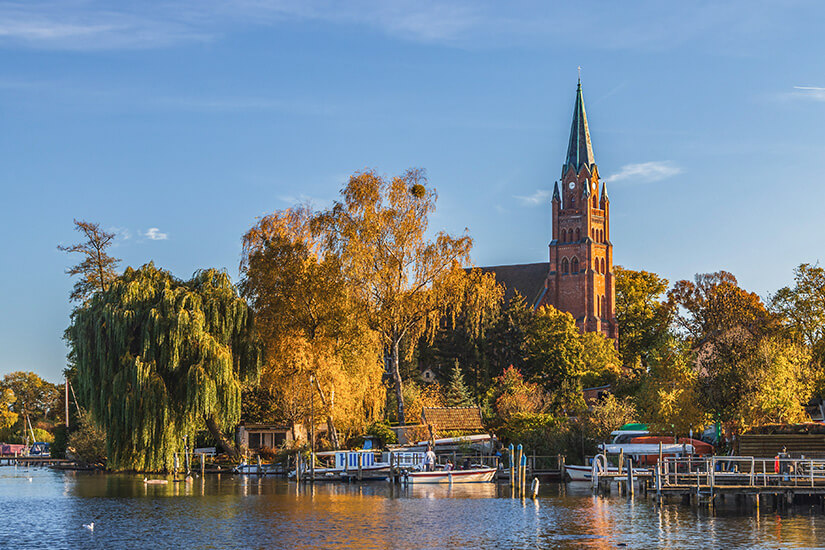 Ein herbstlicher Blick vom Wasser auf das Ufer der Mecklenburgischen Seenplatte. Im Hintergrund ragt eine Backsteinkirche mit spitzem Kirchturm hinter bunt gefärbten Bäumen in Gelb- und Orangetönen empor. Am Gewässer befinden sich Stege mit angedockten Booten sowie eine große Trauerweide am linken Bildrand unter einem sonnigen, blau-weißen Himmel. Urlaub mit Hund Geheimtipps Müritz