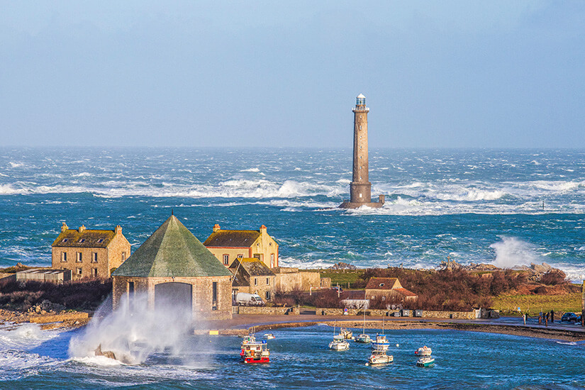Ein hoher, schlanker Leuchtturm ragt mitten aus dem rauen, aufgewühlten Meer vor der Küste der Cotentin-Halbinsel empor. Im Vordergrund befindet sich ein kleiner Hafen mit mehreren verankerten Fischerbooten und historischen Natursteinhäusern am Ufer. Mächtige Meereswellen brechen sich in weißer Gischt an der Hafenmauer unter einem klaren, hellblauen Himmel. Urlaub mit Hund Geheimtipps Contentin Halbinsel