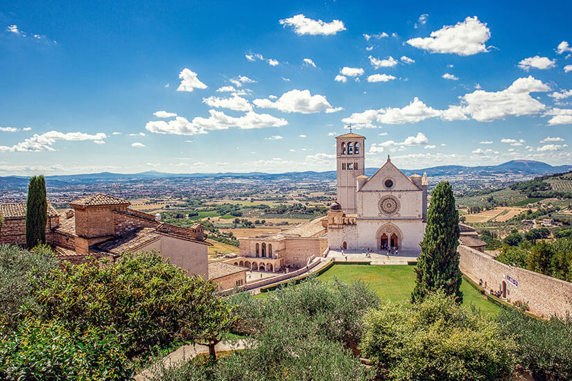 Blick von oben auf die imposante Basilika San Francesco in Assisi in Umbrien. Das helle Steinbauwerk mit großem Rosettenfenster und hohem Glockenturm hebt sich vor einer weitläufigen, grünen Hügellandschaft ab. Im Vordergrund liegen Olivenbäume und dunkle Zypressen, während sich darüber ein strahlend blauer Sommerhimmel mit lockeren Schönwetterwolken erstreckt. Urlaub mit Hund Geheimtipps Umbrien