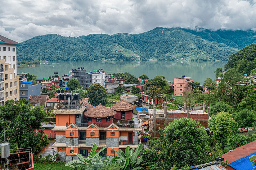 Die Stadt Pokhara mit rötlichen Gebäuden im Grünen ist zu sehen. Im Hintergrund liegt der malerische See. Grüne Hügel spiegeln sich in diesem weider. Nepal Pokhara