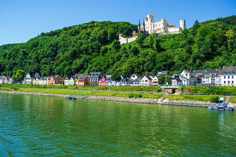 Zu erkennen ist ein breiter Fluss mit einer Uferpromenade, an der sich zahlreiche Häuser entlangziehen. Oberhalb der bewaldeten Hänge thront ein großes Schloss oder eine Burganlage, während der Himmel klar und blau ist. Radfahren Deutschland Rheinradweg