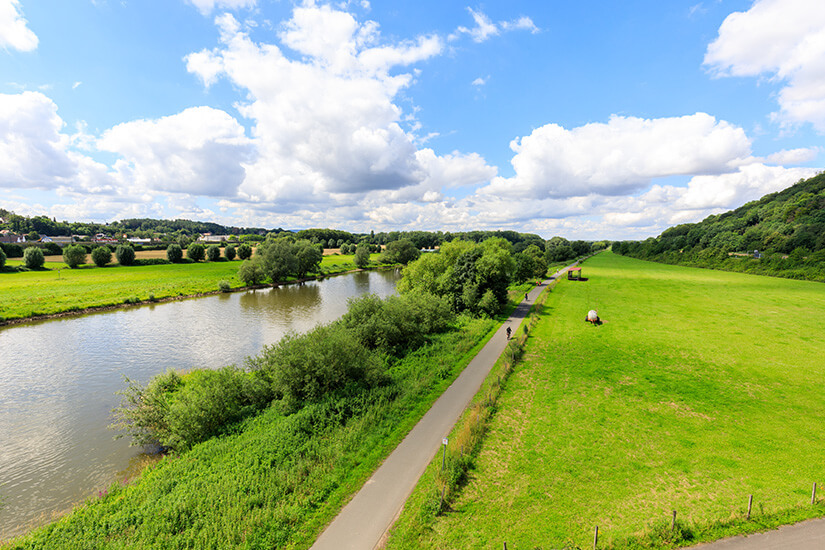 Zu erkennen ist ein Fluss, der sich durch eine weite grüne Landschaft schlängelt, begleitet von einem asphaltierten Radweg. Entlang des Weges sind einzelne Radfahrende unterwegs, während Wiesen, Bäume und ein Himmel mit großen weißen Wolken die Szenerie prägen. Deutschland Weserradweg