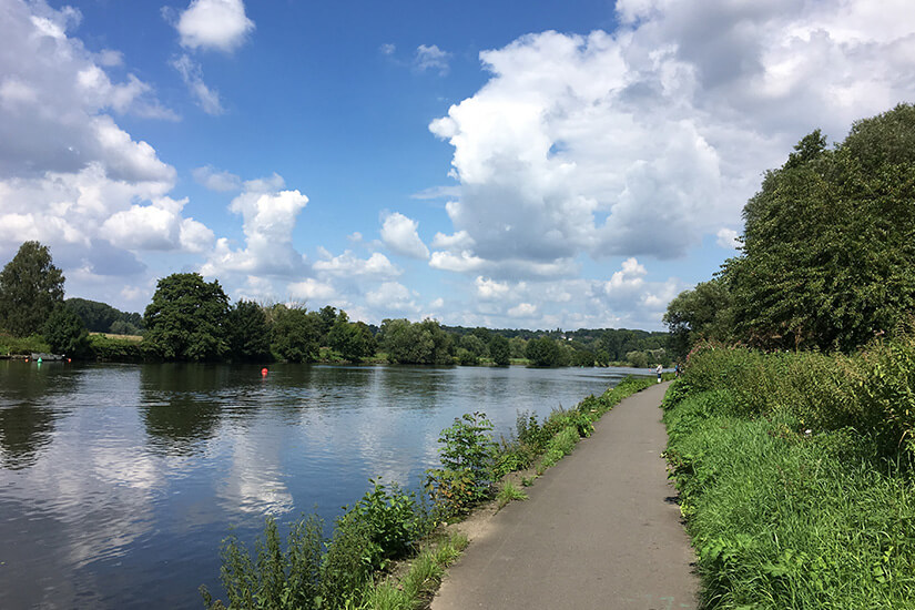 Zu erkennen ist ein asphaltierter Radweg, der direkt entlang eines ruhigen Flusses durch eine grüne Uferlandschaft führt. Dichte Bäume und Sträucher säumen den Weg, während sich darüber ein Himmel mit großen weißen Wolken erstreckt. Deutschland Ruhrtalradweg