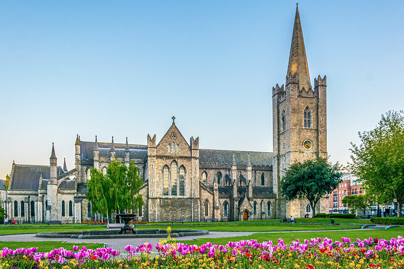 Zu erkennen ist eine große gotische Kathedrale mit einem hohen spitzen Kirchturm, umgeben von einer weitläufigen Grünanlage. Im Vordergrund blühen zahlreiche rosa und gelbe Blumen, während ein Brunnen und gepflegte Rasenflächen die Szenerie ergänzen. Irland Fruehling