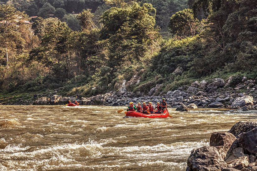 Ein rotes Rafting-Boot mit mehreren Leuten darin fährt über eine Stromschnelle umgeben von grüner Vegetation und Gebirge. Nepal Rafting