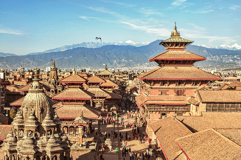 Die roten Dächer einer Stadt in Nepal ragen vor einem Gebirge auf. Ein Vogel fliegt am Himmel. Nepal Anreise