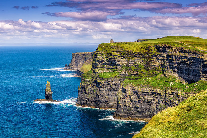Zu erkennen ist eine spektakuläre Steilküste mit schroffen, übereinander geschichteten Felswänden, die steil ins tiefblaue Meer abfallen. Auf den grünen Klippen stehen ein einzelner steinerner Turm und ein markanter Felsen im Wasser, während darüber dramatische Wolken den Himmel prägen. County Clare Cliffs of Moher