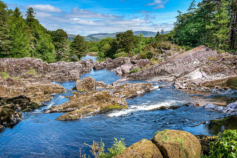 Zu erkennen ist ein klarer Fluss, der sich zwischen großen Felsen und dicht bewaldeten Ufern seinen Weg bahnt. Im Hintergrund erstrecken sich sanfte Hügel und Berge unter einem teilweise bewölkten Himmel. County Kerry Ring of Kerry