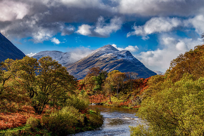 Zu erkennen ist eine Flusslandschaft, die von herbstlich gefärbten Bäumen und dichten Wäldern umgeben ist. Im Hintergrund ragen markante Berge empor, deren Gipfel leicht mit Schnee bedeckt sind, während dunkle Wolken über den Himmel ziehen. Irland Connemara