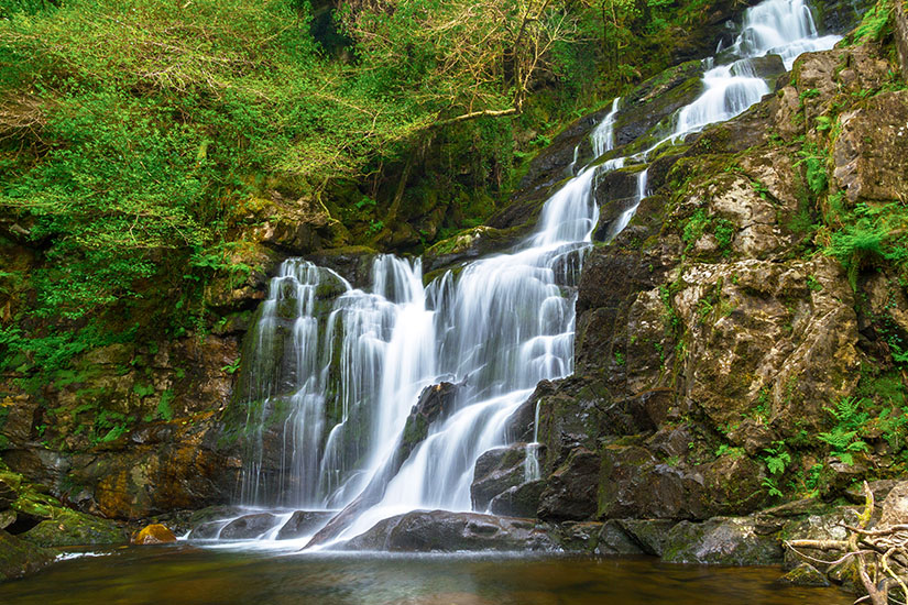Zu erkennen ist ein mehrstufiger Wasserfall, der über dunkle Felsen in ein ruhiges Becken hinabfließt. Dichtes Grün, moosbewachsene Steine und Bäume umgeben die Szenerie und verleihen ihr eine natürliche, idyllische Atmosphäre. Killarney National Park Wasserfall