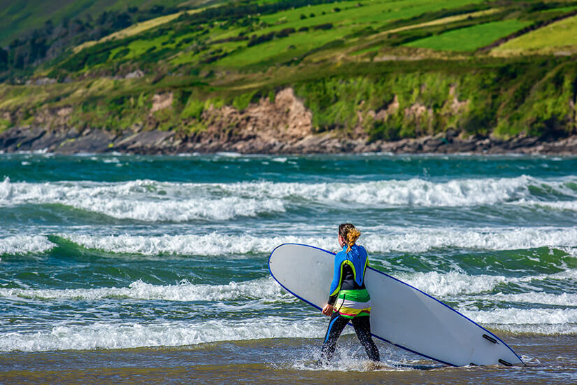 Zu erkennen ist eine Person im Neoprenanzug, die mit einem Surfbrett ins Meer geht, während Wellen auf den Strand treffen. Im Hintergrund erstrecken sich grüne Küstenhänge und felsige Ufer unter einem klaren Himmel. Atlantik Surfen