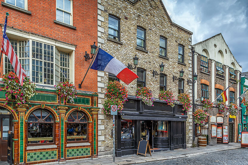 Zu erkennen ist eine Straße mit traditionellen Pubs und historischen Gebäuden, deren Fassaden mit zahlreichen Blumenampeln geschmückt sind. Über den Eingängen hängen verschiedene Nationalflaggen, während Kopfsteinpflaster und bewölkter Himmel das stimmungsvolle Straßenbild ergänzen. Irland Pubs