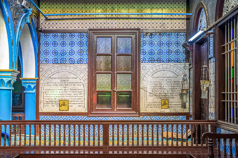 Innenaufnahme der historischen La-Ghriba-Synagoge in Erriadh auf Djerba. Die Wände sind mit blau-weißen Keramikfliesen verziert und tragen Tafeln mit hebräischen Inschriften. Ein Holzfenster mit Butzenscheiben steht im Mittelpunkt, flankiert von blau verzierten Bögen, Säulen und einer hölzernen Balustrade im Vordergrund. Djerba Tunesien La Ghriba