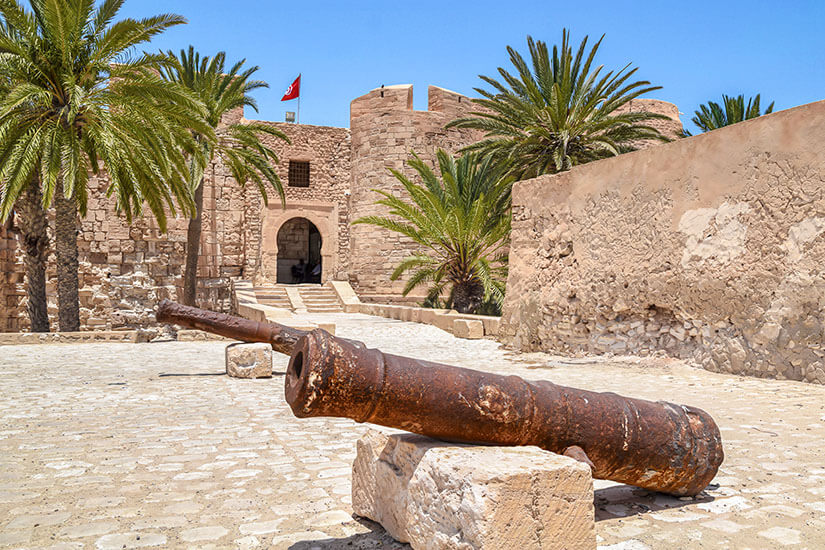 Eine historische Festungsanlage aus hellem Sandstein auf Djerba, auf deren Zinnen die tunesische Flagge weht. Im Vordergrund liegen alte, verrostete Kanonen auf Steinblöcken auf einem gepflasterten Platz. Hohe Dattelpalmen flankieren den rundbogigen Eingang der Festung Borj el Kebir unter einem strahlend blauen Himmel. Djerba Tunesien Borj el Kebir