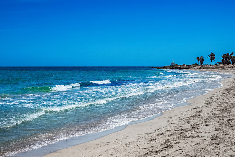 Eine Weitwinkelaufnahme des hellen Sandstrandes an der Blauen Lagune auf Djerba, Tunesien. Das Meer zeigt verschiedene Blau- und Türkistöne mit sanften Wellen. Im Hintergrund säumen Palmen die Küstenlinie, und eine weiße Kuppel ist zu sehen. Ein strahlend blauer, wolkenloser Himmel überspannt die Szene. Djerba Tunesien Blaue Lagune