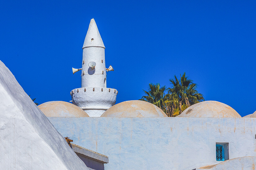 Nahaufnahme der charakteristischen Moschee der Türken in Houmt Souk auf Djerba. Das schneeweiße, spitz zulaufende Minarett mit Lautsprechern hebt sich von sandfarbenen Kuppeldächern ab. Im Hintergrund spitzen grüne Palmen unter einem tiefblauen, strahlenden Himmel hervor. Djerba Tunesien Moschee