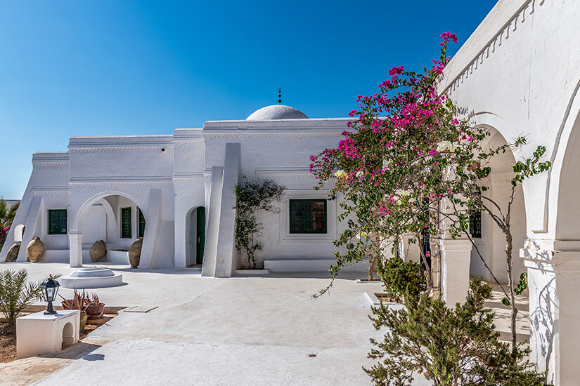 Ein sonniger Innenhof einer traditionellen, schneeweißen Houch-Anlage auf Djerba. Im Vordergrund rankt ein blühender, magenta-farbener Bougainvillea-Strauch. Die weiß getünchten Gebäude mit ihren charakteristischen Bögen, Kuppeln und dunklen Fensterläden stehen unter einem strahlend blauen, wolkenlosen Himmel. Djerba Tunesien Gute Reise