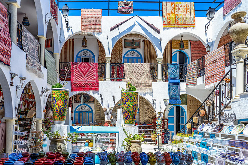 Blick in den farbenfrohen Innenhof eines traditionellen Souks auf Djerba. Über zwei Etagen mit weißen Rundbögen hängen bunt gewobene Teppiche und Wandbehänge über den Geländern. Im Vordergrund und auf Treppenstufen sind handgefertigte Keramiken, bunt bemaltes Geschirr und Keramikvasen übersichtlich aufgereiht. Djerba Tunesien Souvenirs