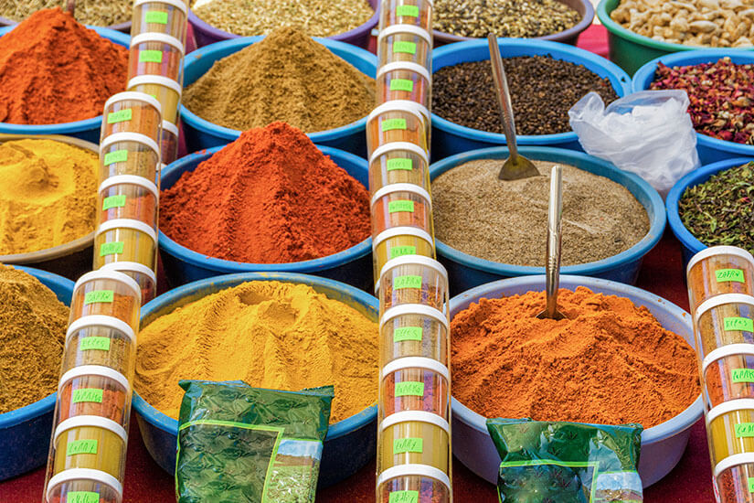 Nahaufnahme von bunten Gewürzbergen auf einem Markt auf Djerba. In blauen und bunten Schalen sind gemahlener Koriander, feuriges Paprikapulver, gelber Kurkuma, Pfefferkörner und getrocknete Blüten aufgetürmt. Zwischen den Schalen stehen durchsichtige Zylinder mit geschichteten Gewürzproben und kleine Probierlöffel stecken in den Gewürzen Djerba Tunesien Gewürze