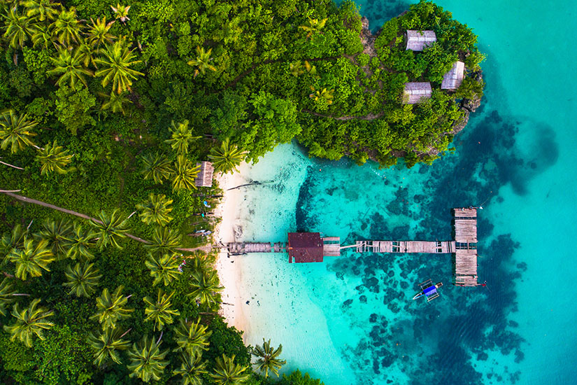 Zu erkennen ist eine tropische Küste mit dichtem Regenwald, Palmen und einem kleinen weißen Sandstrand an türkisblauem Wasser. Ein langer Holzsteg führt ins Meer, während zwischen den Bäumen mehrere einfache Hütten liegen und ein kleines Boot vor der Küste schwimmt. außergewoehnliche reiseziele indonesien