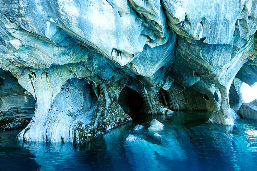 Zu erkennen sind eindrucksvolle, blau schimmernde Fels- oder Marmorhöhlen, deren geschwungene Wände direkt aus dem Wasser aufragen. Das türkisfarbene Wasser spiegelt die außergewöhnlichen Formen und Farbstrukturen der Höhle wider. ausgefallene urlaubsziele chile