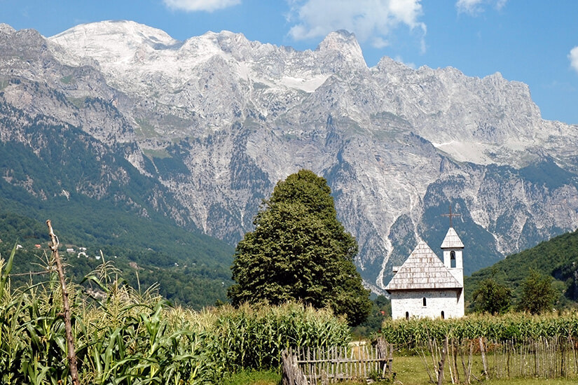 Zu erkennen ist eine kleine weiße Kirche inmitten einer ländlichen Landschaft mit Maisfeldern und einem einfachen Holzzaun. Im Hintergrund erhebt sich ein imposantes, schroffes Gebirgsmassiv unter einem blauen, leicht bewölkten Himmel. interessante urlaubsziele albanien