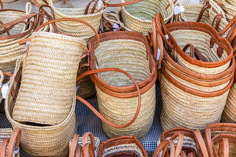 Dicht aneinandergereiht stehen zahlreiche geflochtene Körbe und Taschen aus hellem Palmenblatt mit braunen Ledergriffen und -verstärkungen auf einem Marktstand. Mallorca Souvenirs Llata