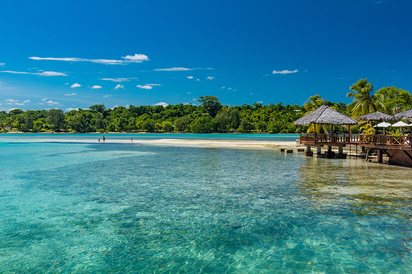 Zu erkennen ist eine tropische Lagune mit kristallklarem, türkisfarbenem Wasser und einem hellen Sandstrand vor dichter grüner Vegetation. Am rechten Ufer stehen strohgedeckte Pavillons auf einem Holzsteg, umgeben von Palmen unter einem tiefblauen Himmel. Tropische Urlaubsziele Vanuatu